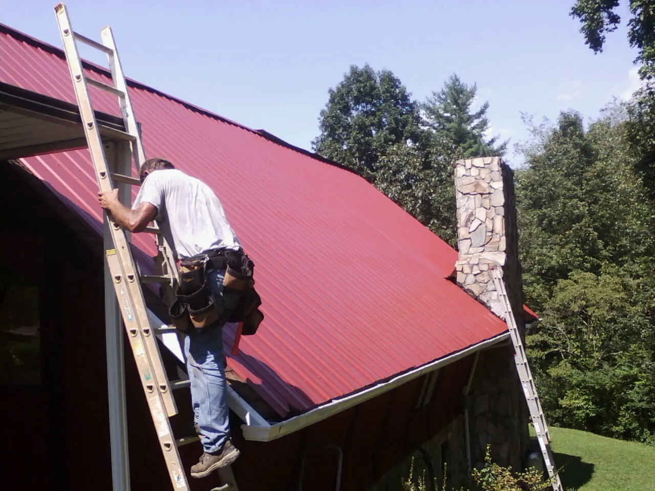 Roofer on a ladder beside a red metal roof and stone chimney — Soffit & Fascia Repair by Roofing Brothers in Keosauqua